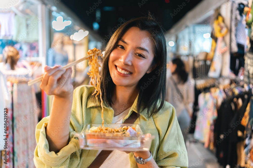 Fototapeta premium Asian woman enjoy eating noodles street food at night market. Traveler Asian blogger women Happy tourists Beautiful female with Traditional thailand bangkok food.