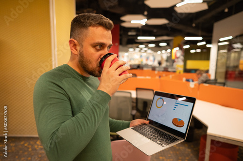 Bearded man holding laptop and drinking coffee in open space office.