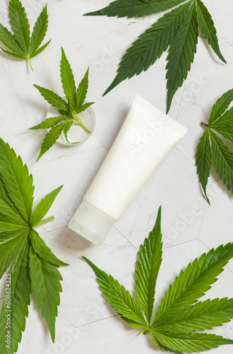 Blank Cream tube near green cannabis leaves on white table, top view. Cosmetic Mockup