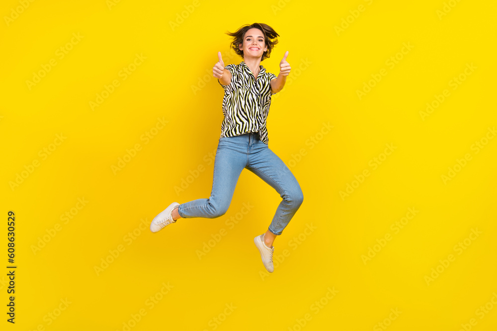 Full length photo of pretty confident woman dressed zebra striped shirt jumping high showing thumbs up isolated yellow color background Full length photo of pretty confident woman dressed zebra striped shirt jumping high showing thumbs up isolated yellow color background