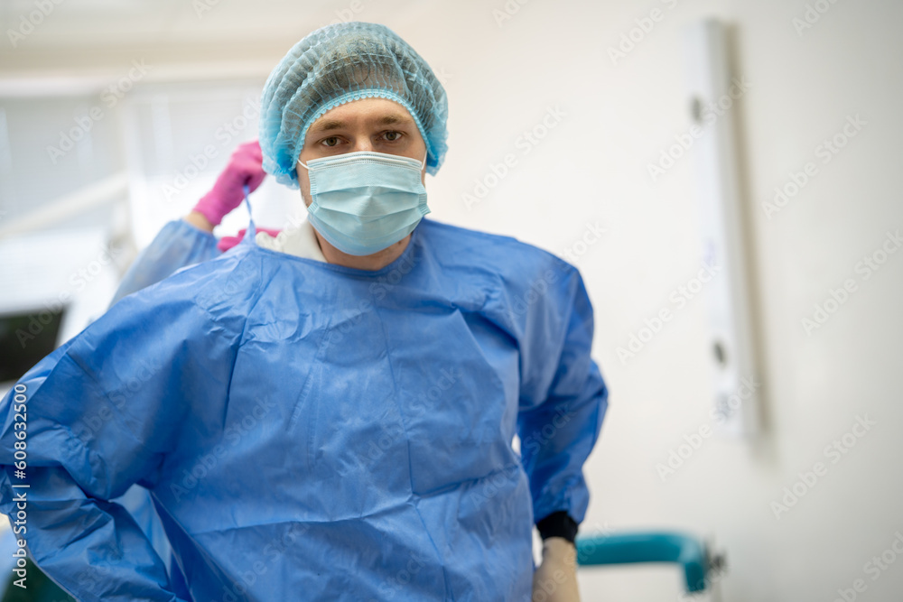 Front view of male dentist in medical uniform wearing blue medical mask and hair cap while unseen assistant adjusts his apron in dental cabinet 