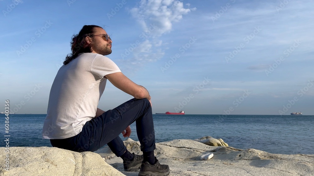 a man in a white t-shirt and glasses sits near the blue sea