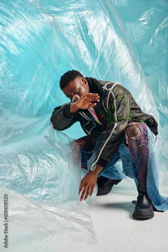 creative expression, DIY clothing, young african american model in outwear jacket with led stripes and trendy jeans posing and covering face near cellophane on turquoise background