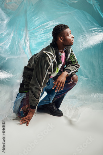 Side view of good looking afroamerican man in outwear jacket with led stripes and ripped jeans looking up near glossy cellophane on turquoise background, creative expression, DIY clothing