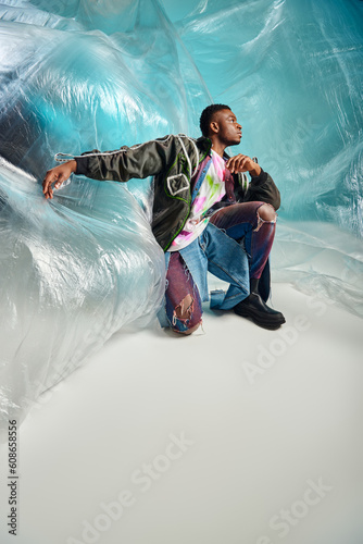 Full length of young afroamerican model in ripped jeans and outwear jacket with led stripes looking away near cellophane on turquoise background, modern pose, creative expression, DIY clothing