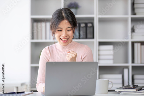 Portrait of a pretty and cheerful Asian businesswoman in casual clothes working at her desk in the modern office, showing her fist, rejoicing after receiving an unexpected email.