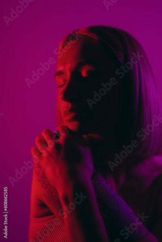 Dreamy female model closing eyes while praying for good against violet background in neon illumination