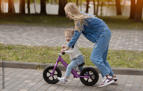 Fototapeta Naklejka Na Ścianę i Meble -  Young mother teaches her little son how to ride a balance bike in the park