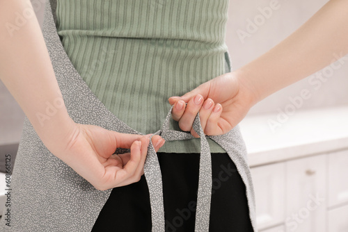 Woman putting on grey apron indoors, closeup