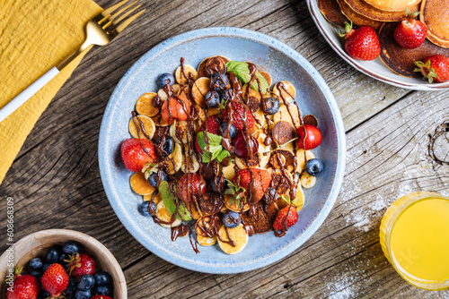 Small American pancakes mixed with seasonal fruit like strawberries, blueberries and cherries. Dipped with melted chocolate, placed on a rustic wooden board. 