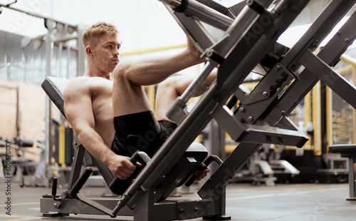 Young muscular man naked torso exercise using press machine to strength his legs in sport gym. Fit man training body workout doing heavy weights leg pushing equipment with quad muscles in fitness club