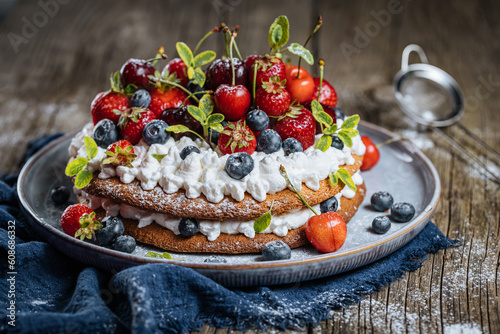 Fresh seasonal layered cake with spring ripe fruit like strawberries, blueberries and cherries. Placed on a elegant blue plate on rustic wooden board