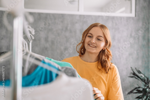 Clothing  purchasing  style concept. Happy blonde haired woman chooses clothes at home wardrobe  holds comfortable jumper on hangers  outfits on racks in background  messages via mobile phone. High