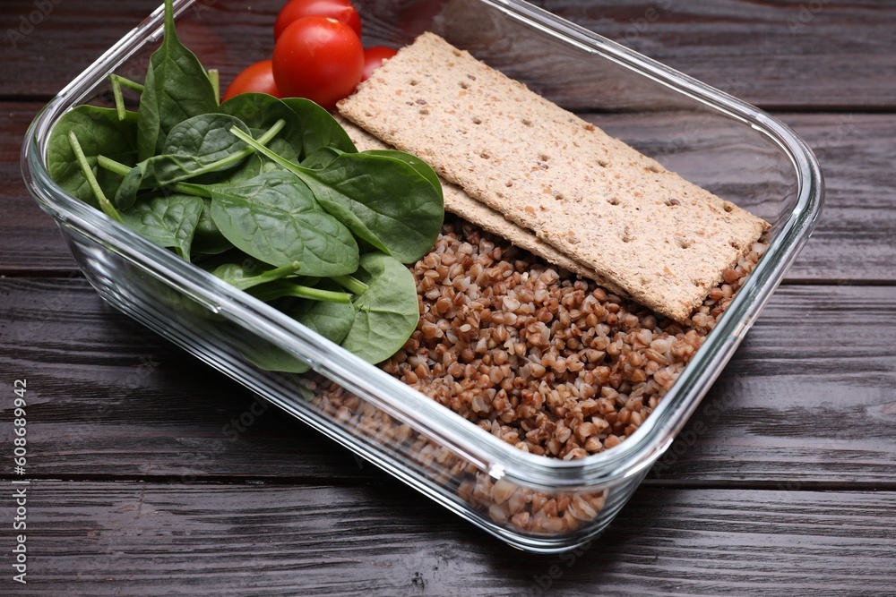 Glass container with buckwheat, fresh tomato, spinach and crispbreads on wooden table, closeup Glass container with buckwheat, fresh tomato, spinach and crispbreads on wooden table, closeup