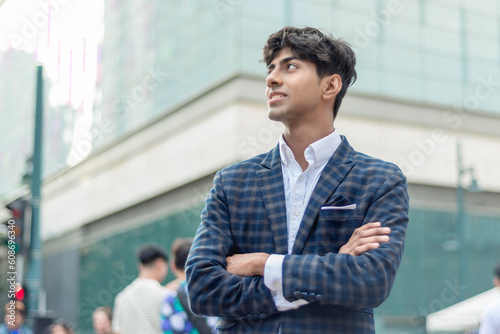 A closer photo of stylish guy in a blue checkered suit, arms crossed, smiling and gazing far away while standing outside, in the city. Building, people and stop light are shown in the background.