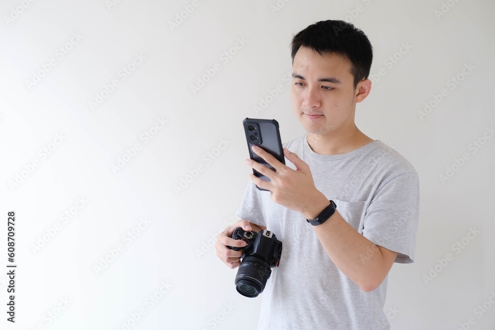 A young Asian man wearing a grey T-shirt is holding and looking at a smartphone in his left hand and holding a DSLR camera in his right hand. Isolated white background. A young Asian man wearing a grey T-shirt is holding and looking at a smartphone in his left hand and holding a DSLR camera in his right hand. Isolated white background.