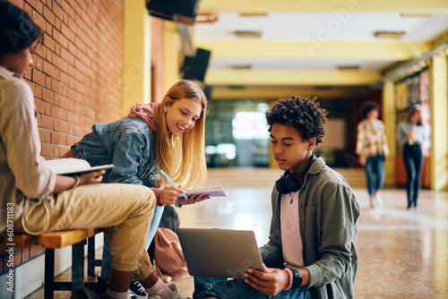 Black teenager using laptop while studying with his friends in high school hallway.