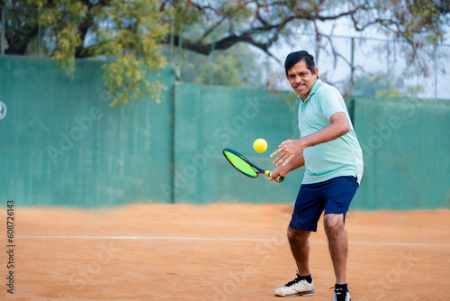 Excited indian senior man busy playing tennis by hitting ball with racquet and court - concept of practicing, healthy lifestyle and sports activity © WESTOCK