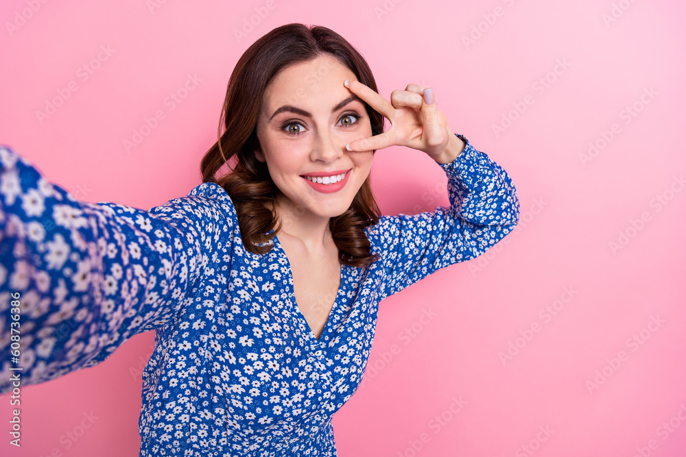 Photo of toothy beaming woman with wavy hairdo wear blue dress making selfie showing v-sign on eye isolated on pink color background Photo of toothy beaming woman with wavy hairdo wear blue dress making selfie showing v-sign on eye isolated on pink color background