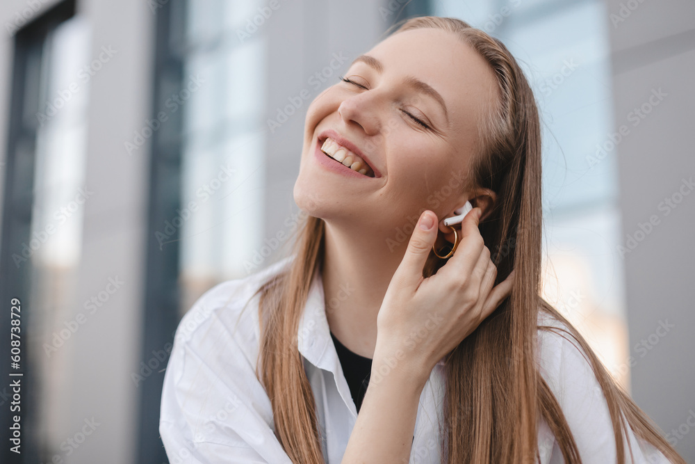 Happy blonde woman sitting on the bench near office building with wireless earbuds in ears and listening music. Smiling girl resting with closed eyes. Use technology, touch headphones.