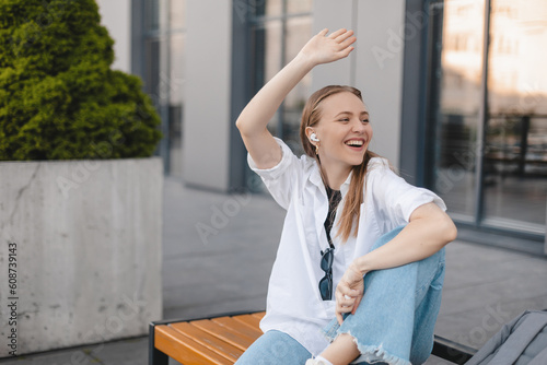 Happy blonde woman make hi gesture, wave her hand while meet colleagues near office building, sitting on bench and listen music in wireless earbuds. Goodbye or hello gesture of business woman. photo