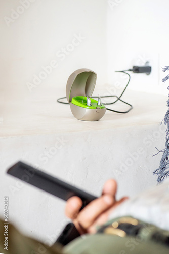 hearing aid for deafness on charger on top of white dining table  detail of unrecognisable eighty year old man s arm and hand on the sofa with the TV remote control.