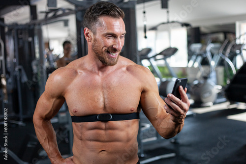 Strong, fit, athletic man in a gym with heart rate monitor around chest checking heart rate on cell phone with a big smile. Good result, good training