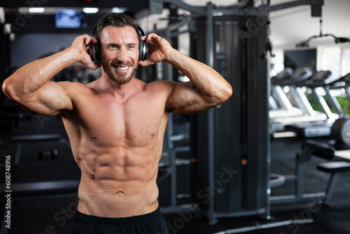 Strong sexy muscular bodybuilder guy standing in a gym with headphones and big smile . Concept gym, crossfit, bodybuilding.