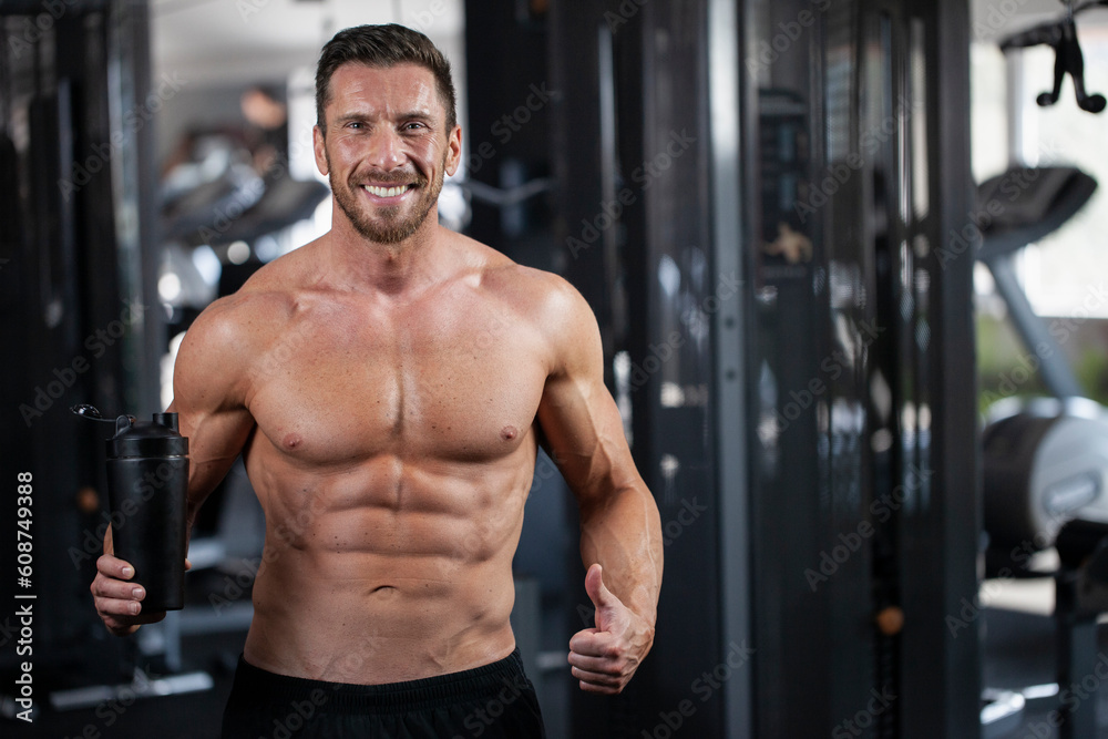 Strong, fit, athletic, attractive man in a gym. Holding black shaker in one hand and shows the thumb up with other hand. Concept protein, water, health. Strong, fit, athletic, attractive man in a gym. Holding black shaker in one hand and shows the thumb up with other hand. Concept protein, water, health.