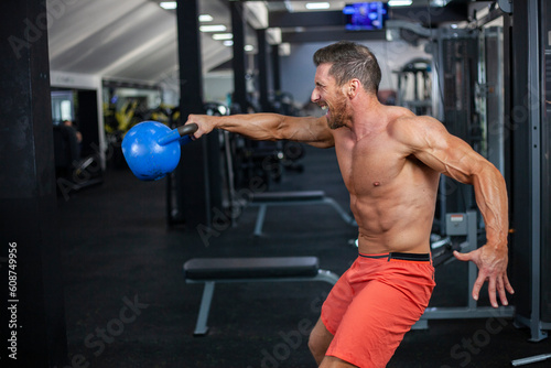 Strong muscular man swings blue kettlebell with one hand in a gym. Powerful and strong face expression