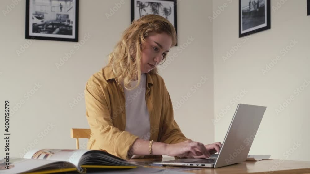 Medium shot of female gallery manager examining printed picture and typing on laptop while working at desk in exhibition hall