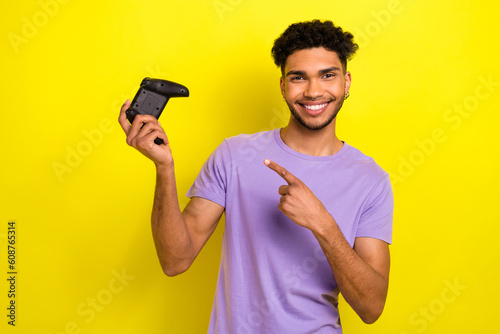 Portrait of handsome cheerful man indicate finger arm hold demonstrate controller isolated on yellow color background