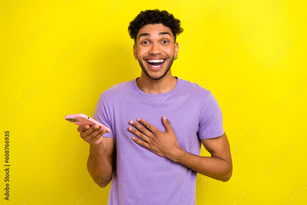 Portrait of astonished handsome man hold smart phone hand touch chest unbelievable news isolated on yellow color background Portrait of astonished handsome man hold smart phone hand touch chest unbelievable news isolated on yellow color background