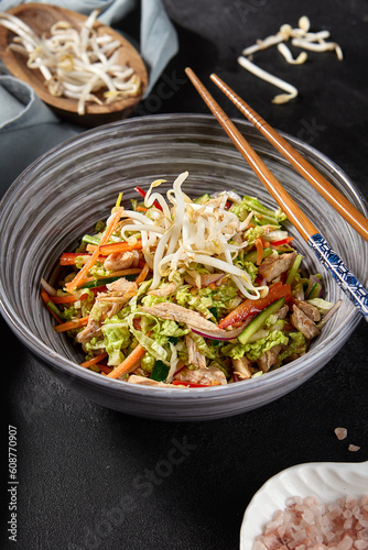 Side view vertical shot of a delightful chicken dish in an Asian style bowl. It's made with vegetables, Peking cabbage, soy sprouts, and a touch of pink sea salt, served on a black