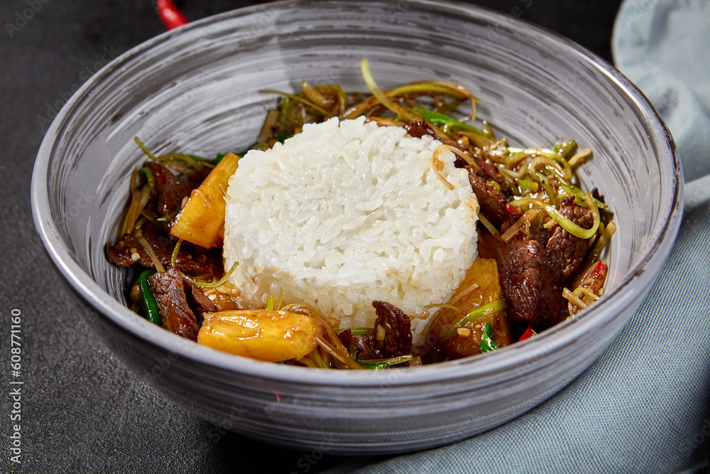 Chinese stir fry with rice and meat with pineapples in a sauce in a gray bowl. Black textured background, fabric, and chili peppers adorn the composition. Side view, vertical frame Chinese stir fry with rice and meat with pineapples in a sauce in a gray bowl. Black textured background, fabric, and chili peppers adorn the composition. Side view, vertical frame