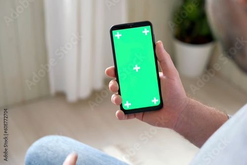 Wallpaper Mural Hipster man sitting at home holding smartphone green mock-up screen in hand. Male person using chroma key mobile phone. Vertical mode. Touching, swiping display, tapping, surfing Internet social media Torontodigital.ca