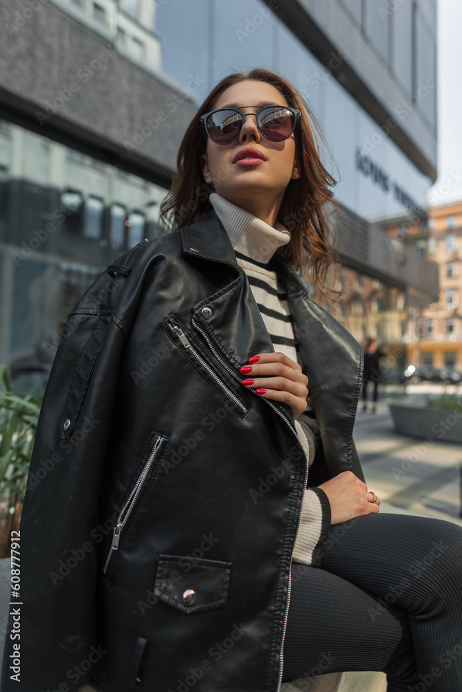 Beautiful fashion young woman model with trendy sunglasses in sweater with black rock leather jacket sits in the city near a modern mall Beautiful fashion young woman model with trendy sunglasses in sweater with black rock leather jacket sits in the city near a modern mall