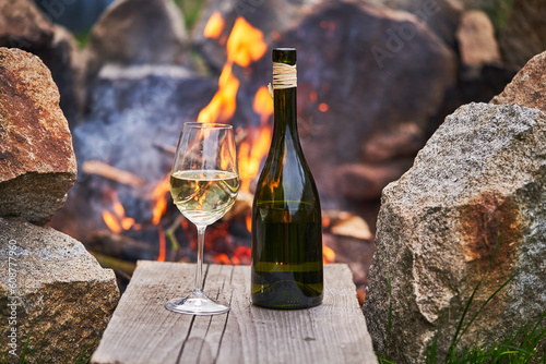 Romantic picture of the glass of white wine and rustic style bottle from small family winery in front of burning stone campfire during the summer sunset in the south moravian region of Czech Republic.