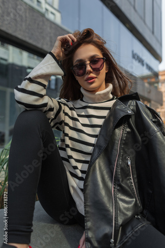 Fashionable young beautiful cool woman with trendy sunglasses in fashion outfit with sweater and black leather jacket sits and poses on the street