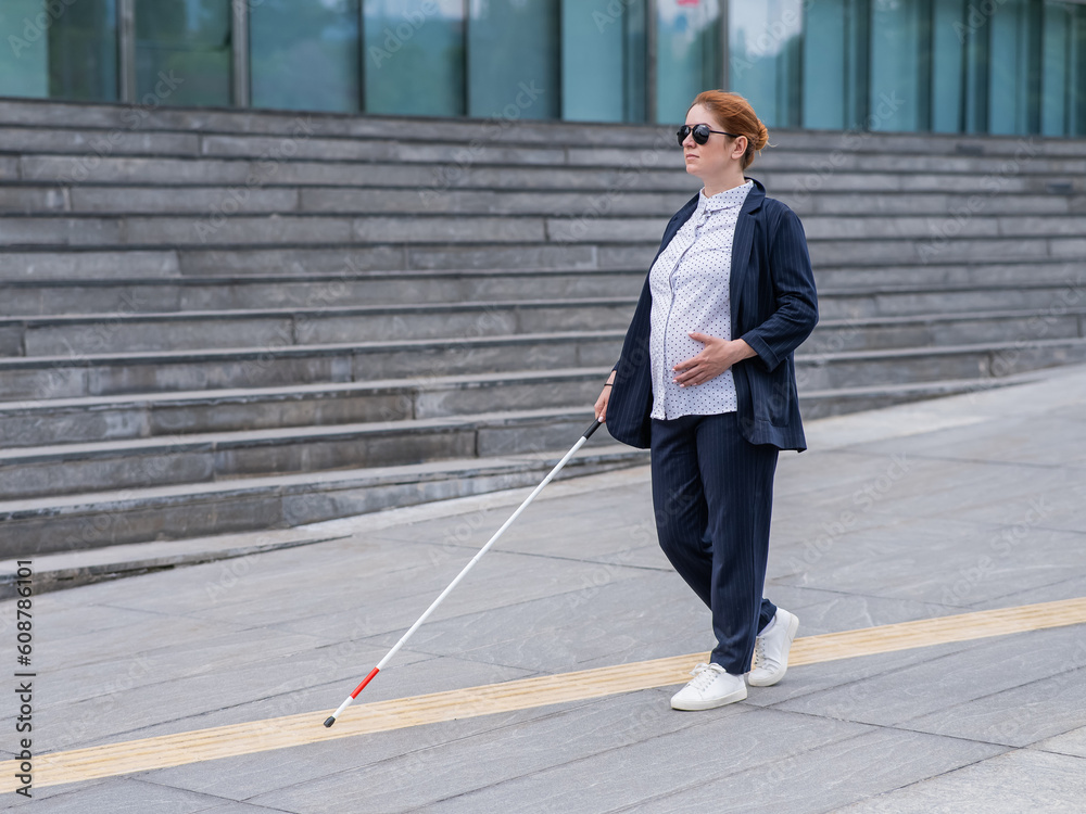 Blind pregnant businesswoman walking along tactile tiles with a cane. 