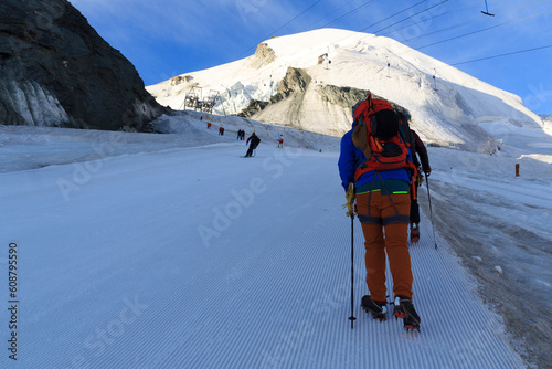 Mountaineer with crampons ascending towards mountain Allalinhorn on ski slope, skiers and snow mountain panorama in Saas-Fee, Pennine Alps, Switzerland