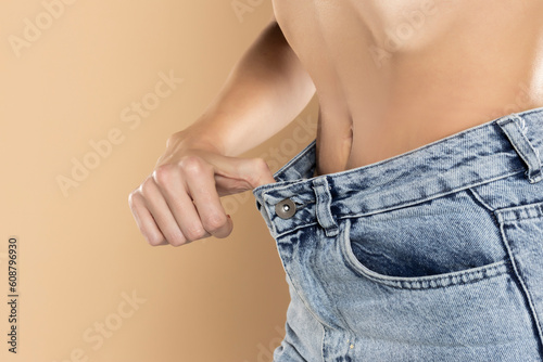 Woman showing slim body after sport trainings, healthy eating. Weight loss concept. Thin woman in big pants,  Slim girl wearing oversized pants on a beige background photo