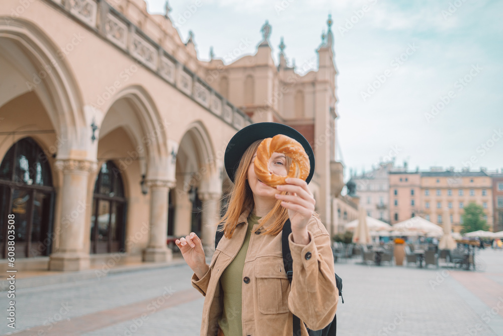 Fototapeta premium Stylish woman tourist holding prezel, traditional polish snack on the Market square in Krakow. Tourist hand holding pretzel on the Cracow Cloth Hall background. High quality photo