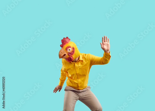 Happy guy in yellow chicken mask having fun at crazy party. Funny cheerful carefree young man wearing bizarre bird mask dancing isolated on turquoise background