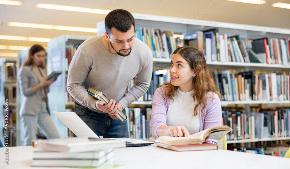 Fototapeta premium Young woman and man working with laptop and books, finding information at library together