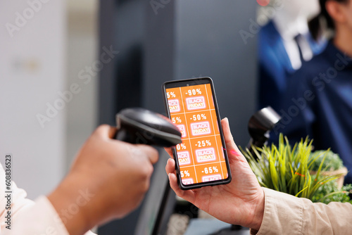 Retail employee scanning smartphone with Black Friday discount coupon while serving customer at cash register desk, close up. Clothing store marketing promotion during seasonal sales