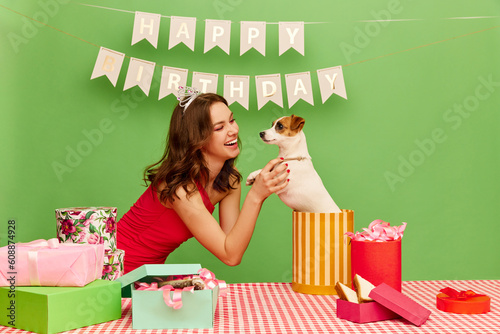 Happy, smiling, young girl celebrating her birthday, opening gift with little dog inside against green background. Best surprise, present. Concept of party, celebration, emotions, female beauty, youth photo