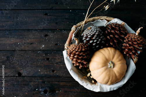 Set of pine cones and holm oak leaves in an esparto basket next to a yellow pumpkin on a wooden table seen from a top view. photo