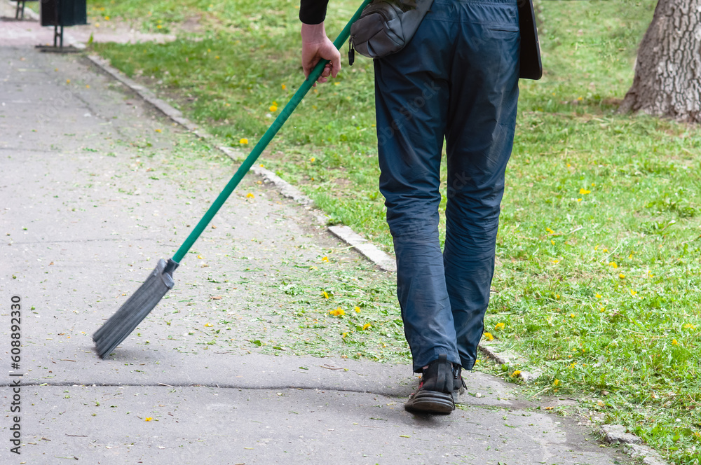 Janitor sweeping a path in an old park on a summer day with a broom. Green cut grass with dandelions. Janitor sweeping a path in an old park on a summer day with a broom. Green cut grass with dandelions.