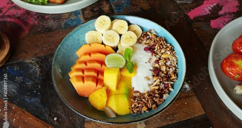 Seasonal tropical fruit salad served in Balinese cafe, sliced fresh fruits served with yogurt and granola on plate. Camera move closer to dish standing on table, selective focus shot, other breakfast photo